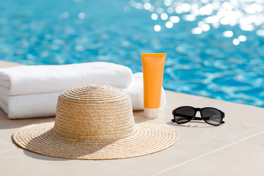 Stylish summer accessories arranged beside a sparkling swimming pool with towel, sunscreen, sunglasses, and woven sun hat