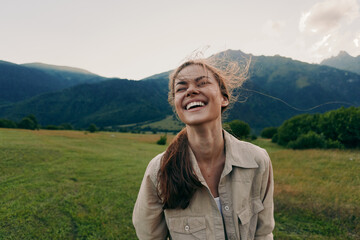 A smiling woman stands in an open field with distant mountains, wearing a beige jacket. The light breeze adds motion for a bright, relaxed lifestyle moment. © SHOTPRIME STUDIO