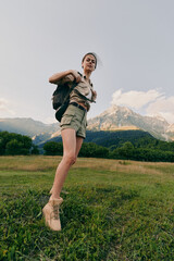 Fototapeta premium Young woman with a backpack leaps across a grassy field as mountains rise behind, capturing a lively outdoor hiking moment under pale blue sky with soft light and a gentle breeze gently.