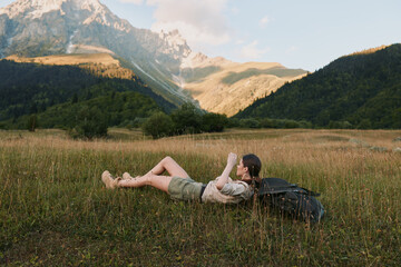 Fototapeta premium Young woman lying on grass in mountain valley reading book during sunny day. Concept of lifestyle, relaxation, and outdoor leisure in nature with backpack nearby.