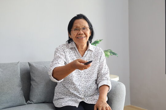 Happy smiling Asian senior woman sitting on couch in the living room, pointing tv remote controller to switch channels, watching favorite movie online series