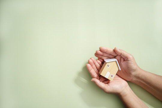 Hands hold wooden house model on light green background. Real estate, buy and sale, Property insurance, property protection, real estate safety, and residential care.