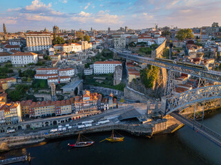 Naklejka premium Drone view of Dom Luis I Bridge over Douro River in Porto, Portugal, with Gaia waterfront, including a tram on the bridge and rabelo boats by the quay at sunrise