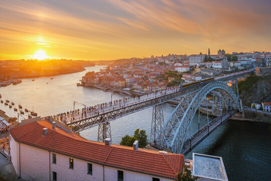 Drone view of Dom Luis I Bridge over Douro River in Porto, Portugal, with Ribeira waterfront, including a tram on the bridge and rabelo boats by the quay at sunset