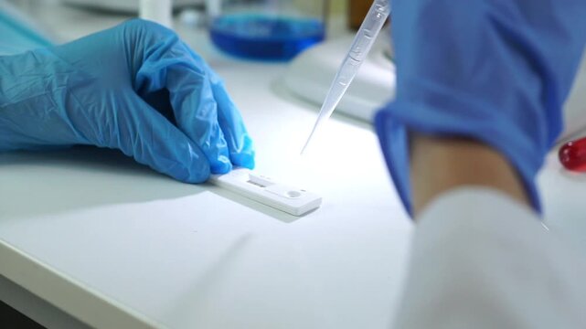 Close-up of a researcher in blue gloves using a pipette to drop a liquid sample onto a rapid test cassette for diagnostic analysis.