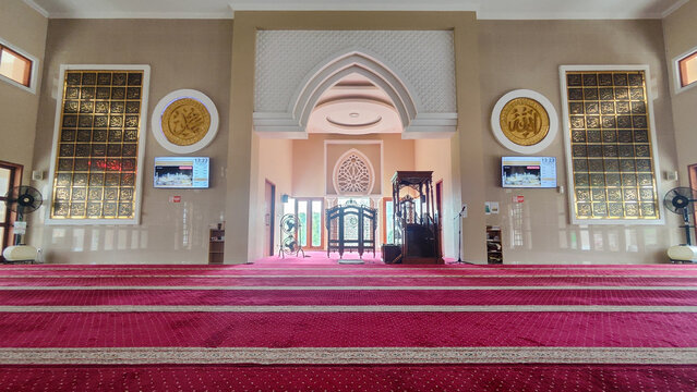 ​A wide interior view of a mosque featuring a red prayer carpet, a decorative mihrab, and golden Islamic calligraphy on the walls.