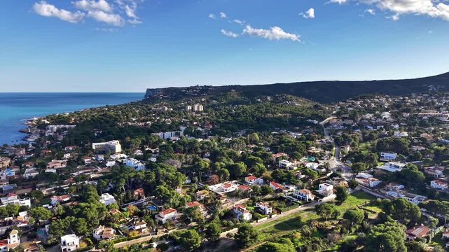 Sunlit coastal village aerial view, turquoise sea and cliff backdrop, rows of white villas and lush gardens,