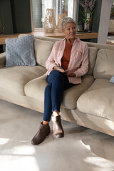Naklejka premium Mature adult woman sitting on beige sectional sofa holding smartphone wearing pink overshirt