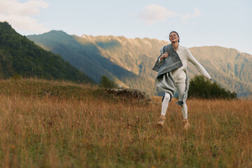 Fototapeta premium Young woman with long hair joyfully runs through a grassy field in casual clothes during golden hour with mountains in the background and a relaxed expression on her face.