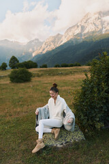 Fototapeta premium Woman outdoors in a mountain meadow, wearing a cozy sweater, sitting on a rock, relaxed pose, soft natural light, enjoying the fresh breeze, lifestyle moment