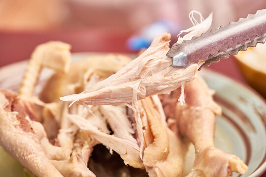 A macro shot of Baeksuk, traditional Korean boiled chicken. Metal tongs lift a piece of breast meat, highlighting the dense, fibrous texture of native chicken, capturing the authentic taste of Korea.