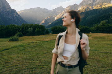 Fototapeta premium Young woman with short hair enjoys a mountain hike wearing casual clothes and earbuds while standing in a green meadow with forest and peaks in the background.