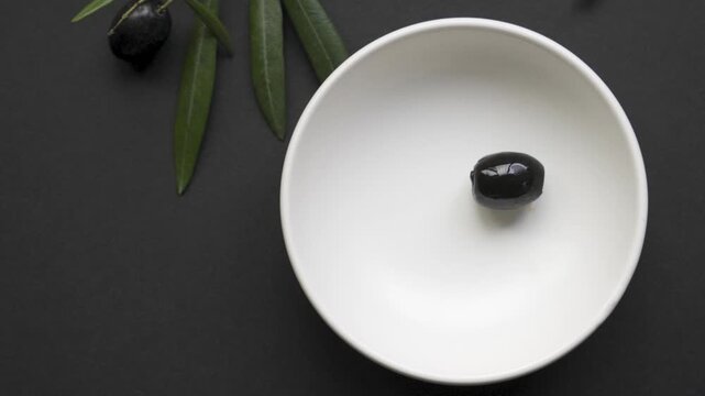 Black_olives_placed_with_tweezers_into_empty_white_bowl_on_black_background_4k_120fps