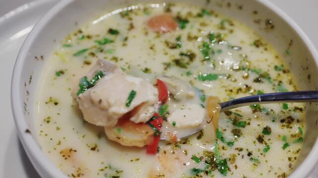 Spoon lifting piece of chicken from white cream soup with herbs and vegetables in ceramic bowl clip.