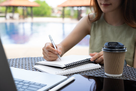 Woman writing in notebook with laptop and coffee cup on table, remote work at outdoors cafe. digital nomad lifestyle, online business concept
