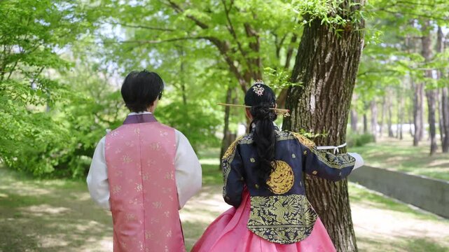 Two figures in traditional Korean attire stroll through a sunlit park. The woman wears a vibrant pink skirt with gold embroidery. Her companion dons a matching vest over a white tunic