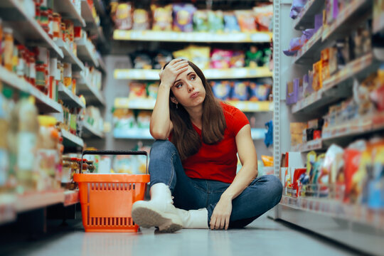 Exhausted Female Shopper Sitting On Floor With Empty Basket Outdoors. Frustrated woman feeling stressed out while looking at supermarket shelves