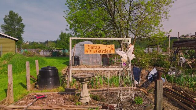Reversing low altitude perspective pulls back from a personalized community garden plot featuring a custom wooden sign engraved with Saras Garden. The eclectic space contains a stone birdbath white