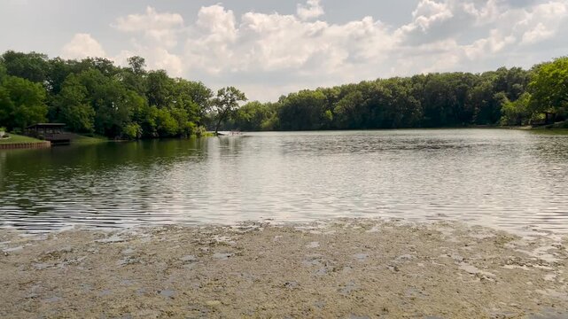 Horizontal pan across the lake at Weldon Springs State Park, Illinois. The smooth shot reveals lush shoreline greenery and the Veterans Point Memorial under a bright summer sky.