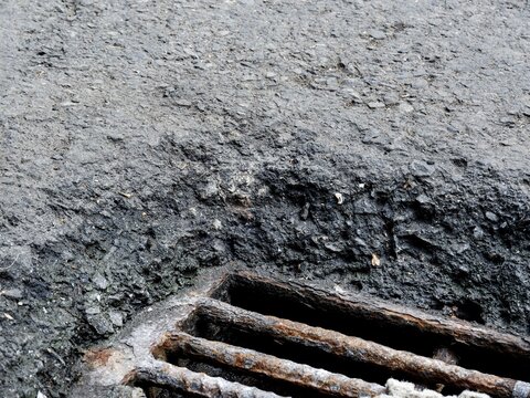 A high-angle close-up showing the thick, layered edge of broken asphalt pavement surrounding a rusted iron drainage grate on an urban street.