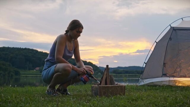 Woman crouching on grass using butane gas torch to ignite campfire near tent and calm lake at sunset, capturing outdoor camping activity and peaceful natural atmosphere in Savci Slovenia