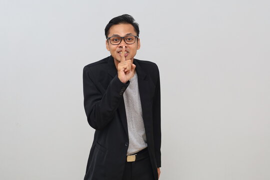 Young male businessman smiling while making a wordless gesture, pressing a finger to his lips, hiding a secret, a secret concept, standing in front of a white background.