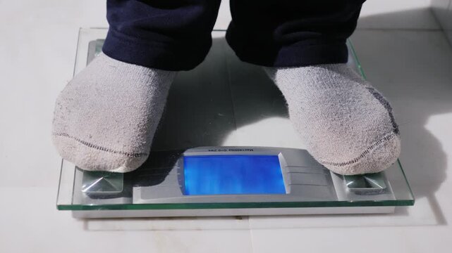 A close up motion shot of a person stepping on a digital scale to check weight on a white floor, displaying the numbers on the screen.