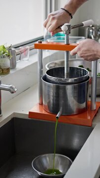 Close-up of hands operating a manual press juicer, extracting vibrant green juice into a bowl over a kitchen sink. Fresh liquid streams steadily from the spout, vertical