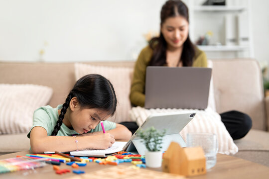 Asian mother working on laptop while daughter doing homework at home