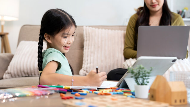 Smiling Asian girl homeschooling with tablet while mother working behind