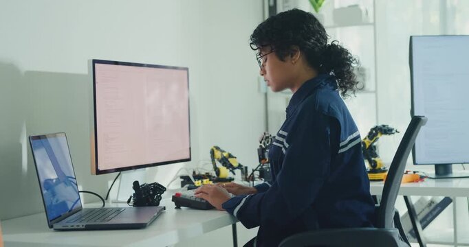 Young African American female robotics engineer coding AI software for robotic arm in laboratory. Embedded system, firmware testing, machine learning, industrial automation, future technology concept.