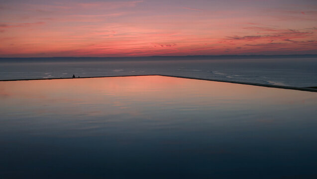 Infinity edge pool with reflection of pink sunset sky over calm ocean horizon, luxury resort coastal escape with peaceful tranquil atmosphere