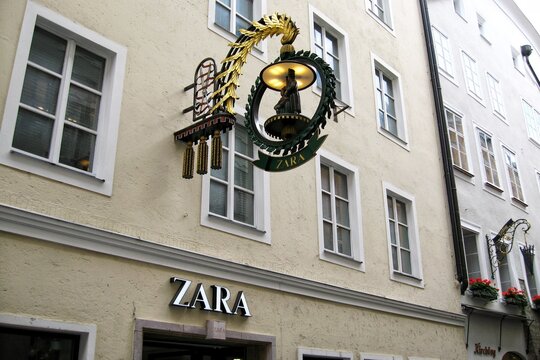 Ornate wrought iron guild sign with a figure and decorative gold elements  of Zara shop hanging from a historic light yellow building facade on a narrow street. Salzburg, Austria - August 14, 2009