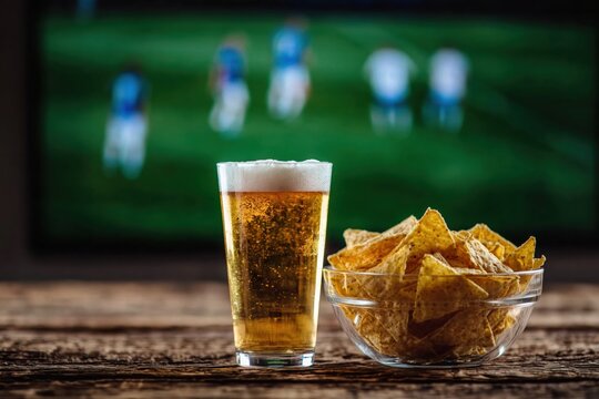 Glass of refreshing lager beer next to bowl of crunchy potato chips with blurry soccer match playing on television screen in background. High quality photo