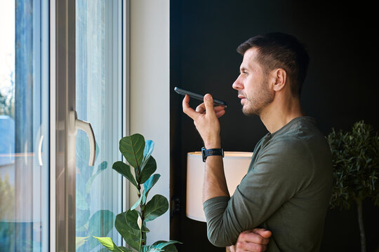 Man speaks into smartphone while standing by window at home. Concept of voice messaging and mobile communication.