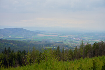 Obraz premium Wide view from a mountain slope over green forest, rolling hills, farmland and small villages in a misty valley under cloudy sky. Nature, rural landscape, travel, countryside and environment concepts