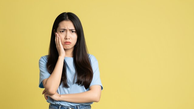 Expression of Doubt: A young woman expresses doubt, with a contemplative expression. Her hand touches her jawline, reflecting deep thought and uncertainty, against a simple backdrop.