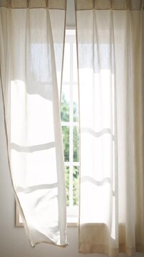 Sheer Beige Curtains Blowing in Sunlight by a White Window