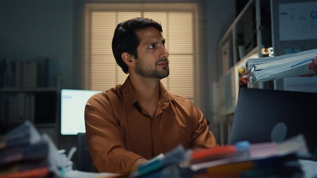 Overworked Latin businessman receiving extra paperwork from boss at desk in dark night office. Working late overload, deadline burnout, heavy overtime stress, corporate business problem concept.