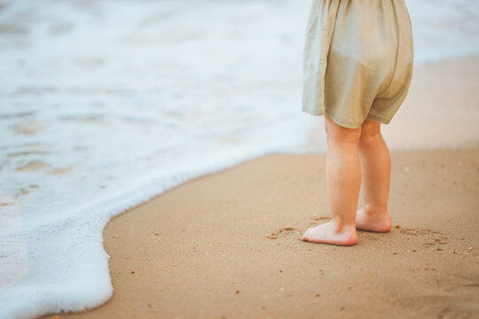 Little Asian toddler girl standing barefoot on a sandy beach watching sea waves during a summer family vacation. Close-up of child's feet on the shore. Concept of childhood discovery, nature