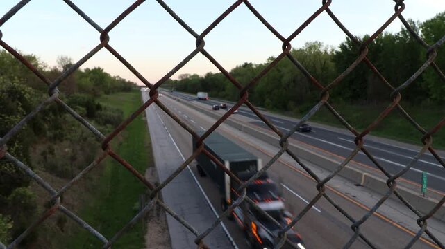 A metal rusted chain link fence in focus with cars and semi trucks driving down the highway road blurred out of focus behind the fence. 