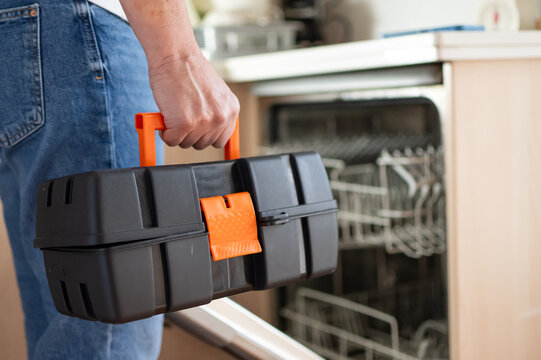 Hand carrying a black toolbox with orange accents towards an open dishwasher, indicating readiness for home maintenance or diy appliance repair in a kitchen