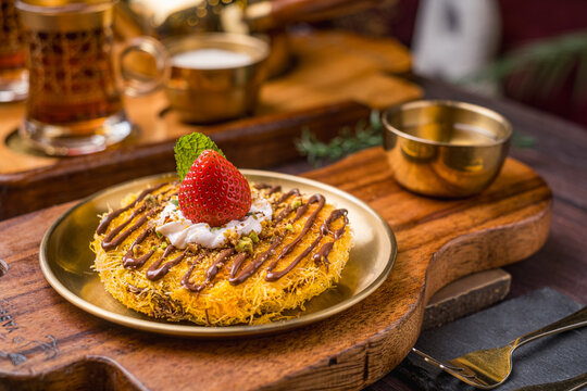 kunafa served on a plate and topped with chocolate jam, whipped cream, strawberries and chopped pistachios. Arranged on a wooden plate.