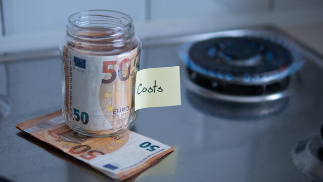 Glass jar with euro banknotes and sticky note reading costs near burning gas stove. Concept of rising energy prices, household expenses, inflation, utility bills and European energy crisis.