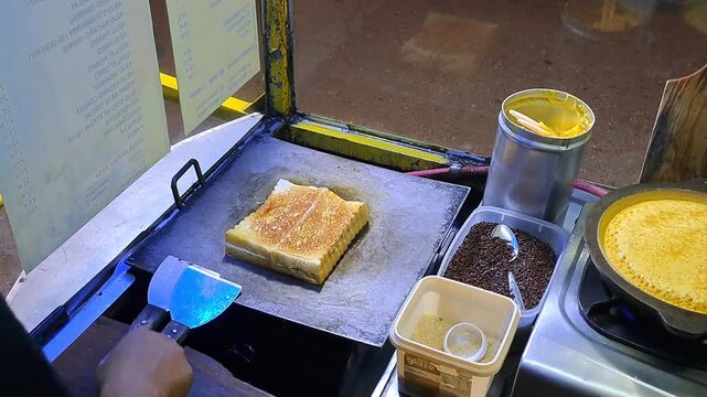 Close-up of a street food vendor preparing authentic Indonesian sweet martabak (Terang Bulan) and grilled toast (Roti Bakar). The footage showcases the detailed process of pouring batter, give butter.