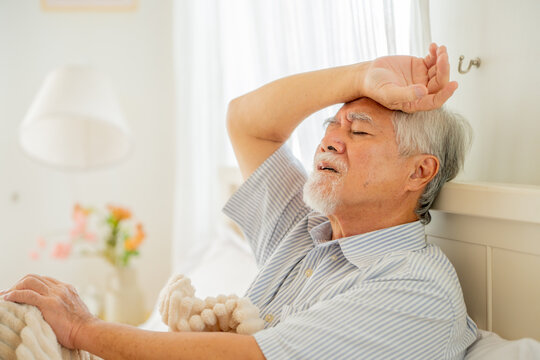 Sick senior Asian man suffering from fever and fatigue in bed at home, elderly male patient feeling unwell with flu or viral infection symptoms, exhausted older adult resting during illness recovery
