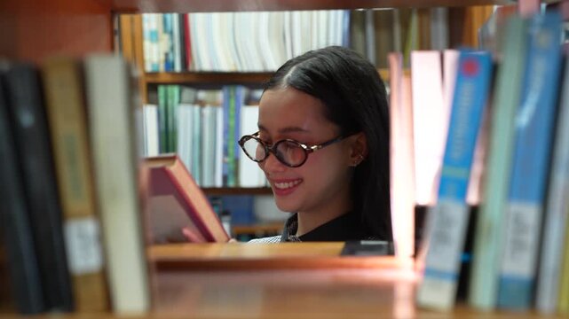 Looking through bookshelves. Asian woman student walking to pick up and read a book in library. Female college student with glasses smiling while studying in university. Education research in school