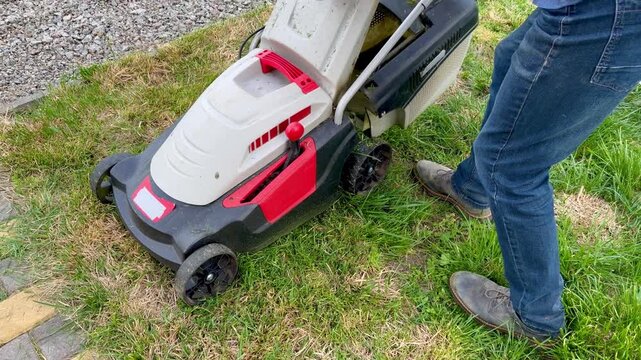 A man in jeans and boots installs a plastic grass catcher box onto a red and white electric lawnmower on a green lawn.