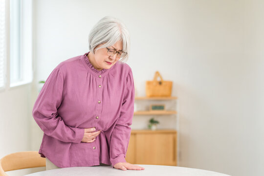 A senior woman with gray hair, clutching her stomach in pain, has a pained expression on her face.