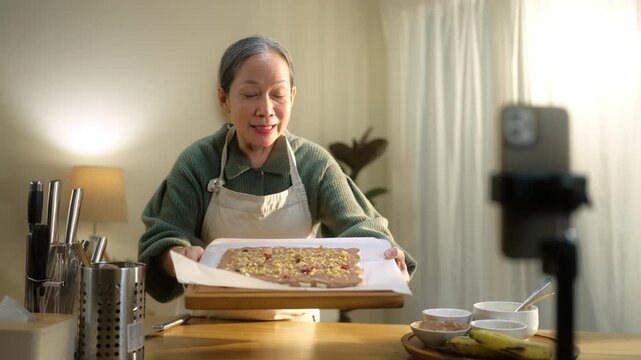 Senior woman livestreams cooking from home on smartphone, highlighting active aging and digital lifestyle. Reflects social media, content creation, creativity, and senior lifestyle.